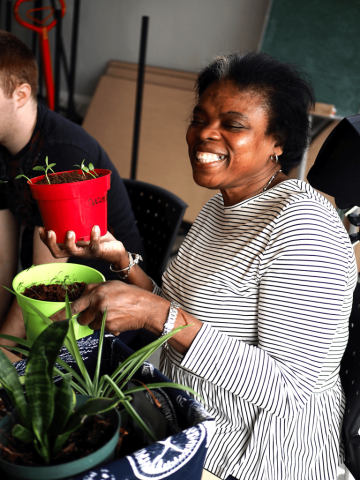 A woman is holding two potted plants and smiling. There is a third potted plant beside her.