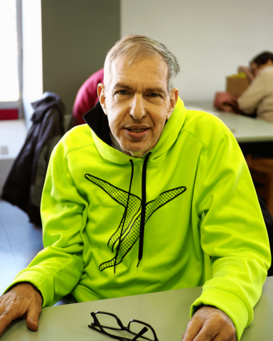 An older man is seated at a desk. He is smiling and wearing a bright sweatshirt.