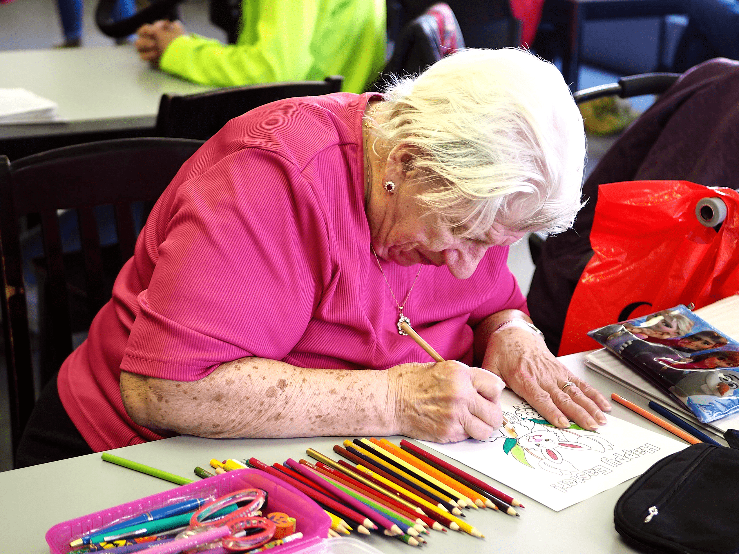 An older woman is seated at a desk colouring a picture. There are many pencil crayons on the desk beside her.
