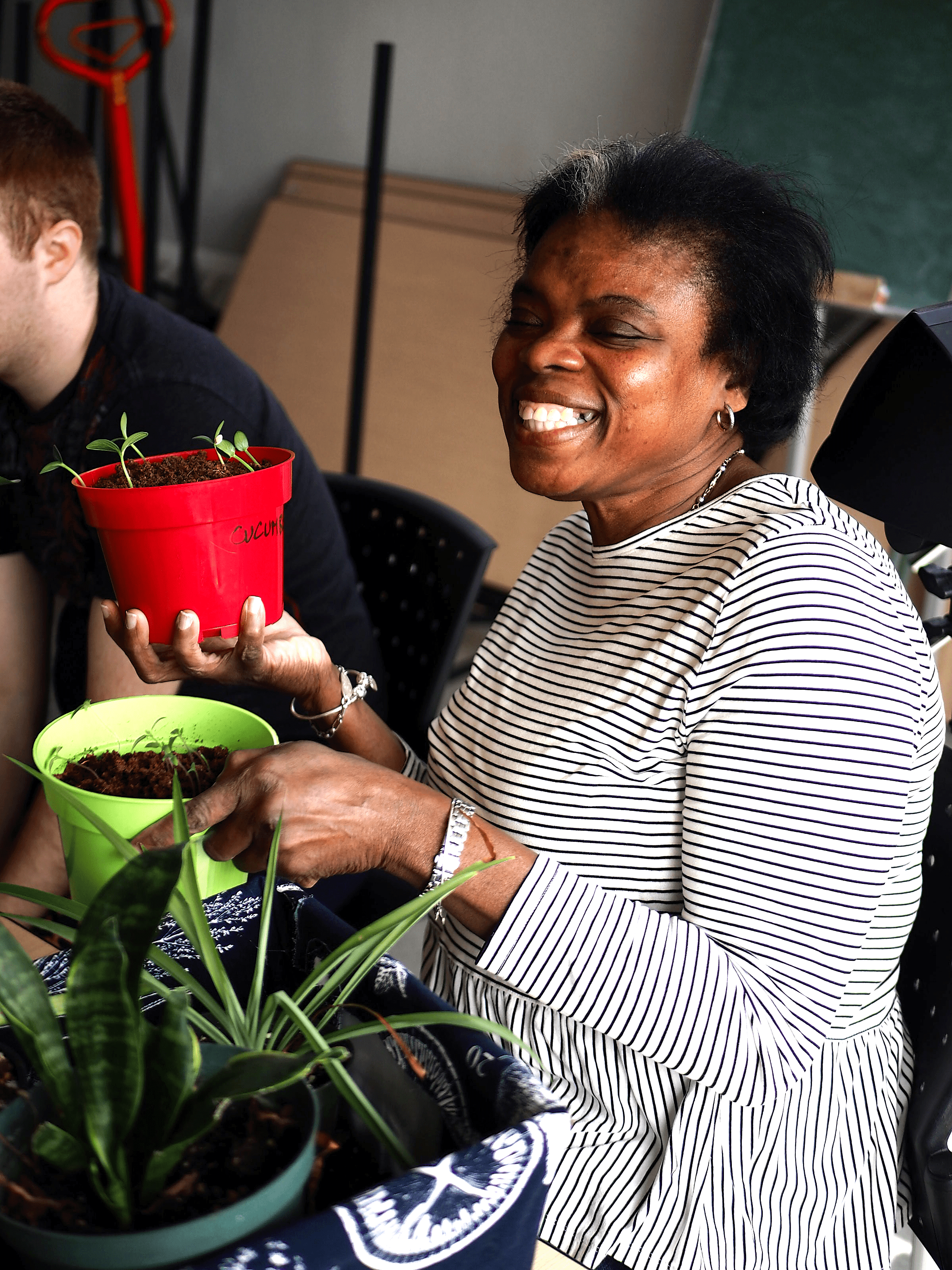 A woman is holding two potted plants and smiling. There is a third potted plant beside her.