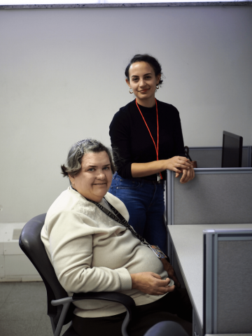 A woman is seated at a computer desk and smiling. Another woman is standing beside her.