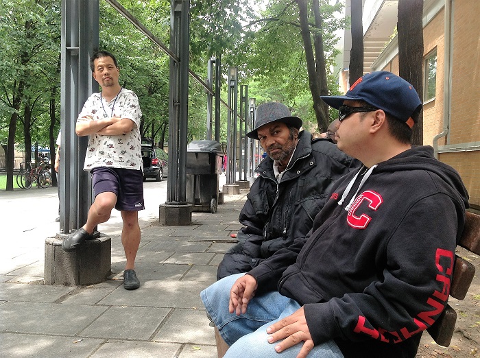 Three men are outside. Two are seated on a bench and one is standing against a column.